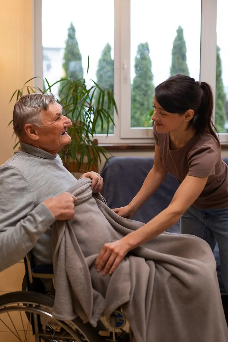 A smiling elderly man in a wheelchair is being covered with a blanket by a younger woman in a room with a view of trees outside