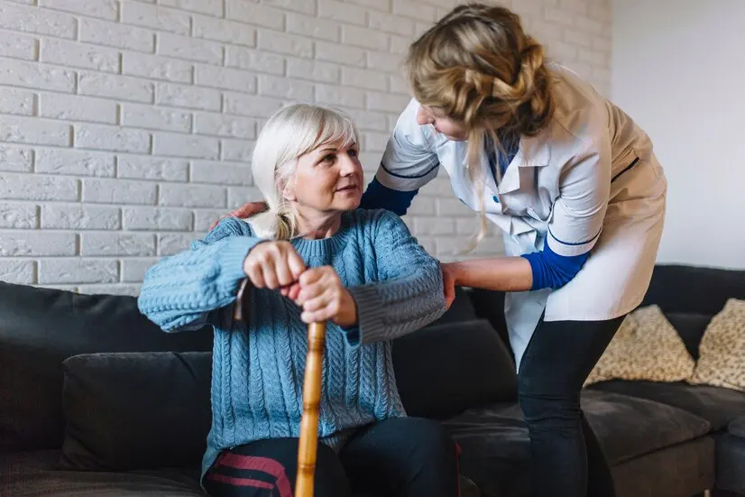 Elderly woman on a sofa with a cane, assisted by a younger woman in a living room